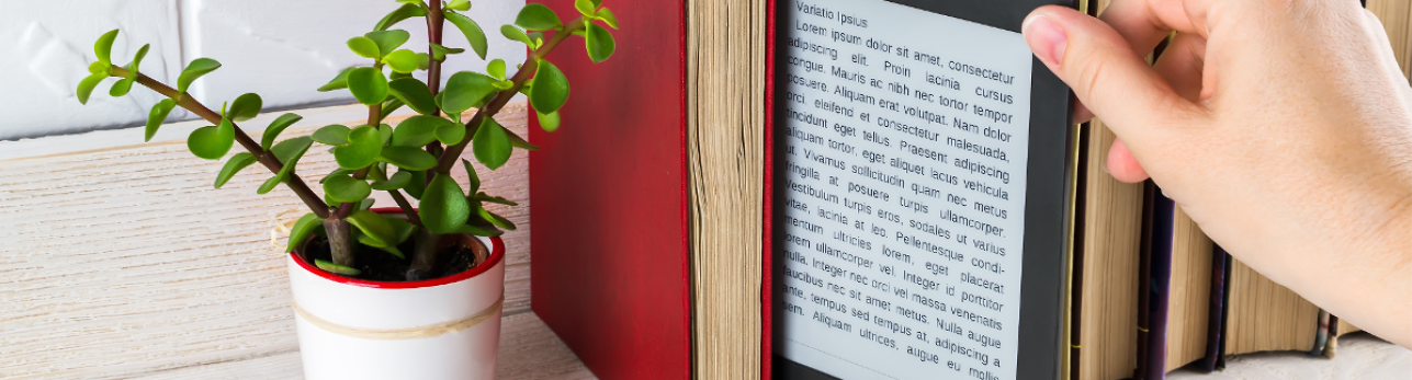 A hand holds an e-reader displaying text between books on a shelf. A small potted plant with green leaves adds a touch of nature to the scene.