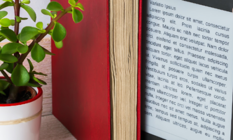 A person's hand holding an e-reader displaying text, surrounded by books and a small potted plant with green leaves, against a wooden surface.